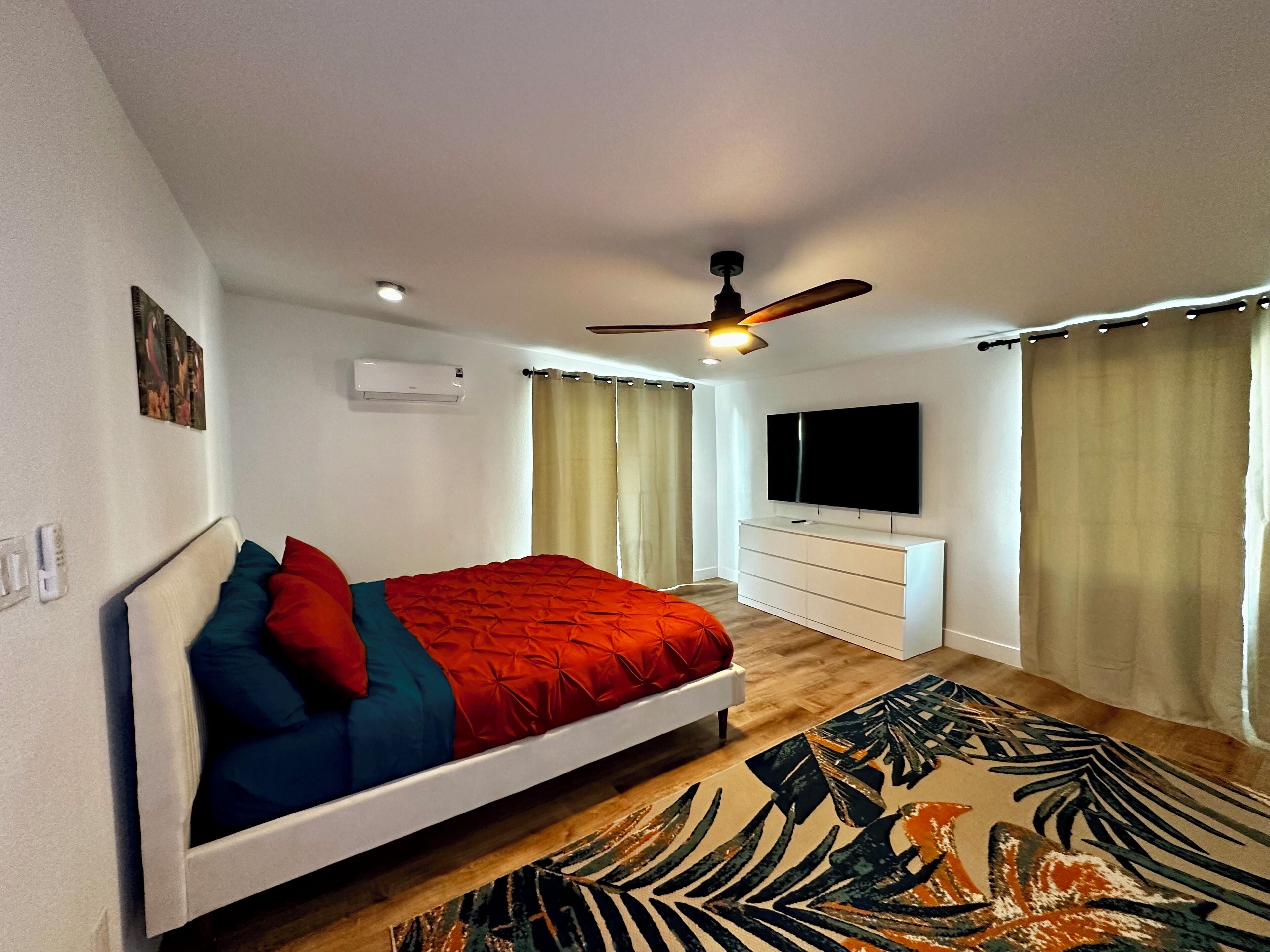 Un dormitorio con cama, ventilador de techo, televisor, cómoda y ventana con cortinas.