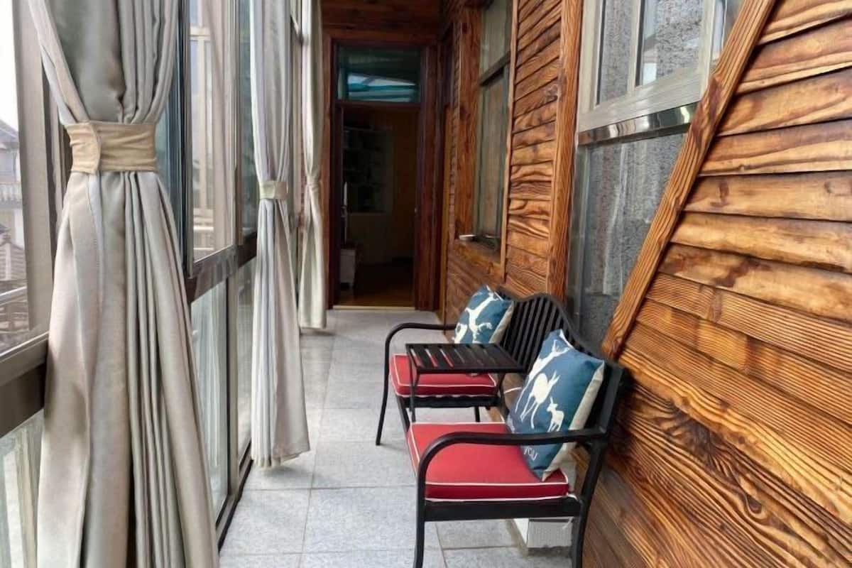 A wooden exterior with two black metal chairs with red cushions and patterned pillows, a wooden door, and a window with curtains.