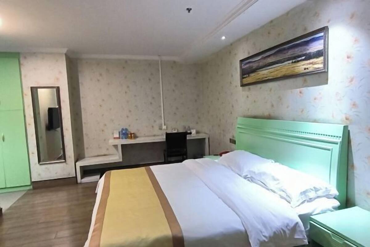 A hotel room with a bed, a bedside table, a desk with a chair, a mirror, and a painting on the wall.