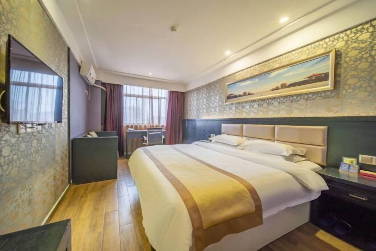 A hotel room with a large bed, a flat-screen TV, a painting on the wall, and a view through the window.