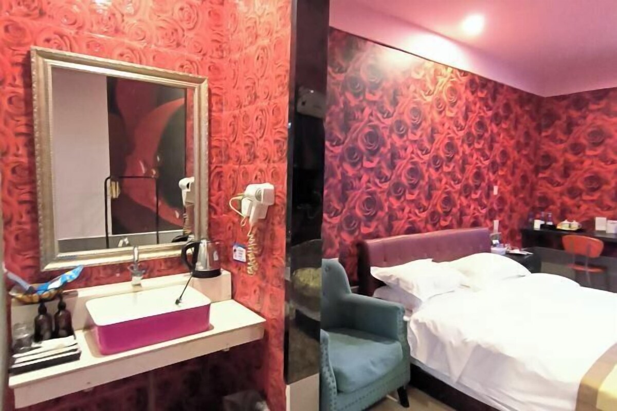 A hotel room with a large mirror, a sink, a bed, a chair, and a red patterned wall.