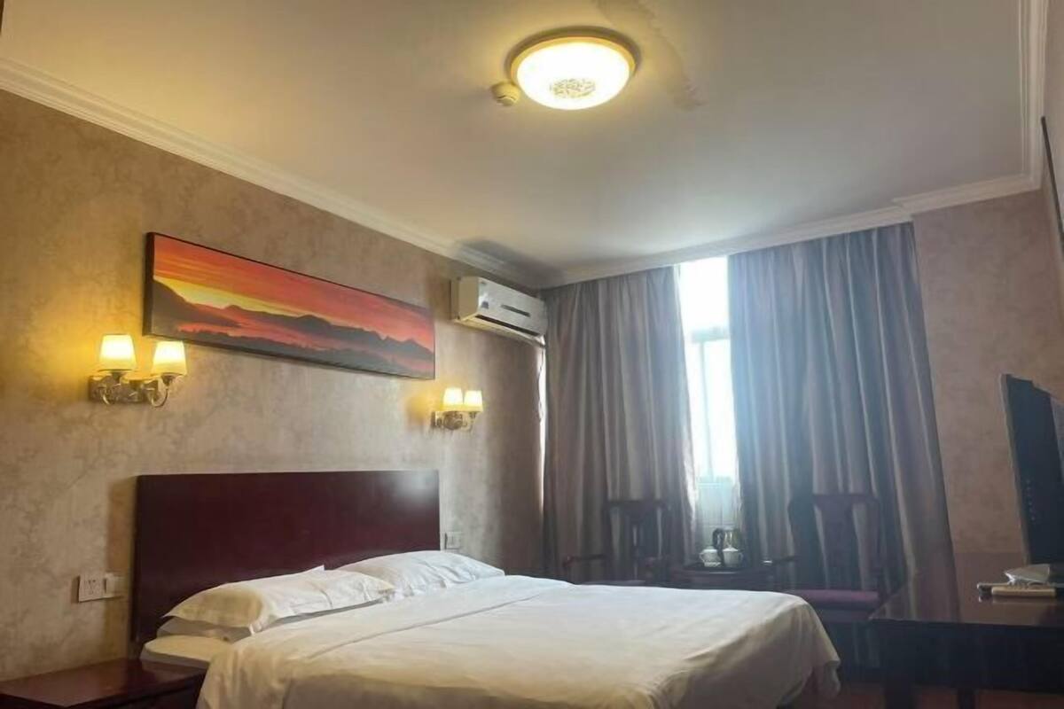 A hotel room with a bed, two wall-mounted lamps, an air conditioning unit, a painting, and a window with curtains.