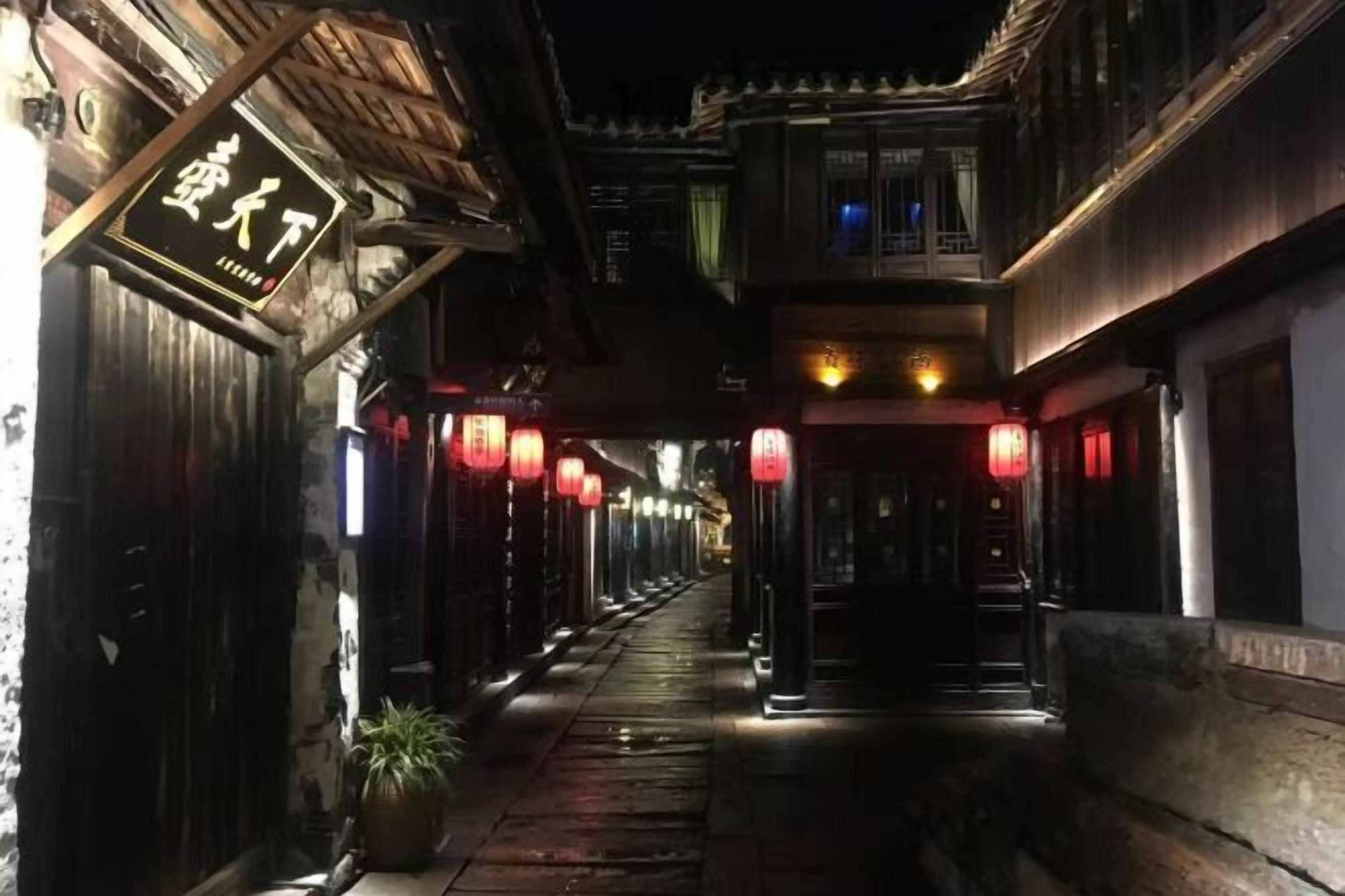 Een smalle steeg met traditionele architectuur, verlicht door rode lantaarns, en een bordje met een aanduiding van een locatie.