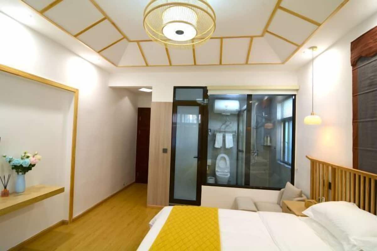 Een hotelkamer met een bed, een nachtkastje met een bloevas, een houten hoofdbord en uitzicht op de badkamer door een glazen deur.