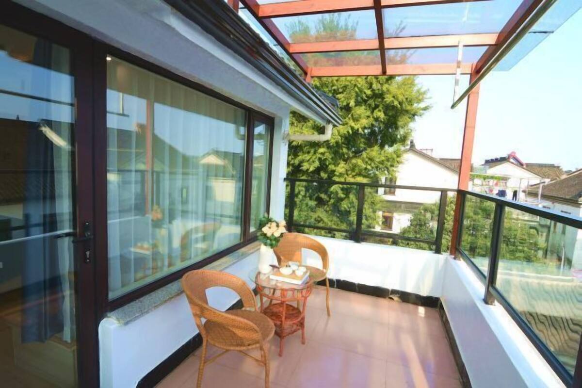 Een balkon met een glazen deur, rieten stoelen, een tafeltje met een vaas en uitzicht op groen en gebouwen.
