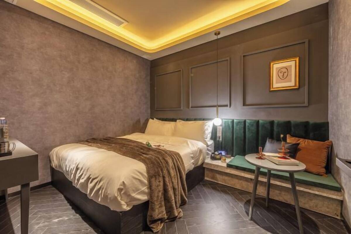 A modern hotel room with a bed, bedside table, lamp, and a seating area with a table and cushions.
