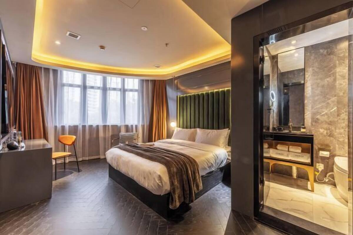 A modern hotel room with a bed, a desk, a chair, and a bathroom visible through a glass partition.