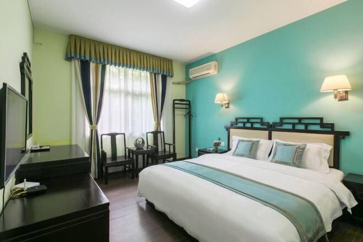 Quarto de hotel com cama, mesas de cabeceira, escrivaninha, cadeira, televisão e janela com cortinas.