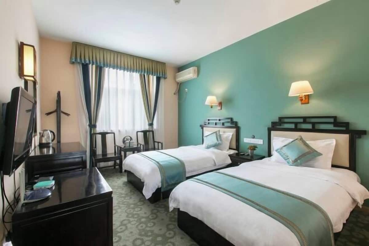 Quarto de hotel com duas camas, uma secretária com televisão, uma cadeira e uma janela com cortinas.
