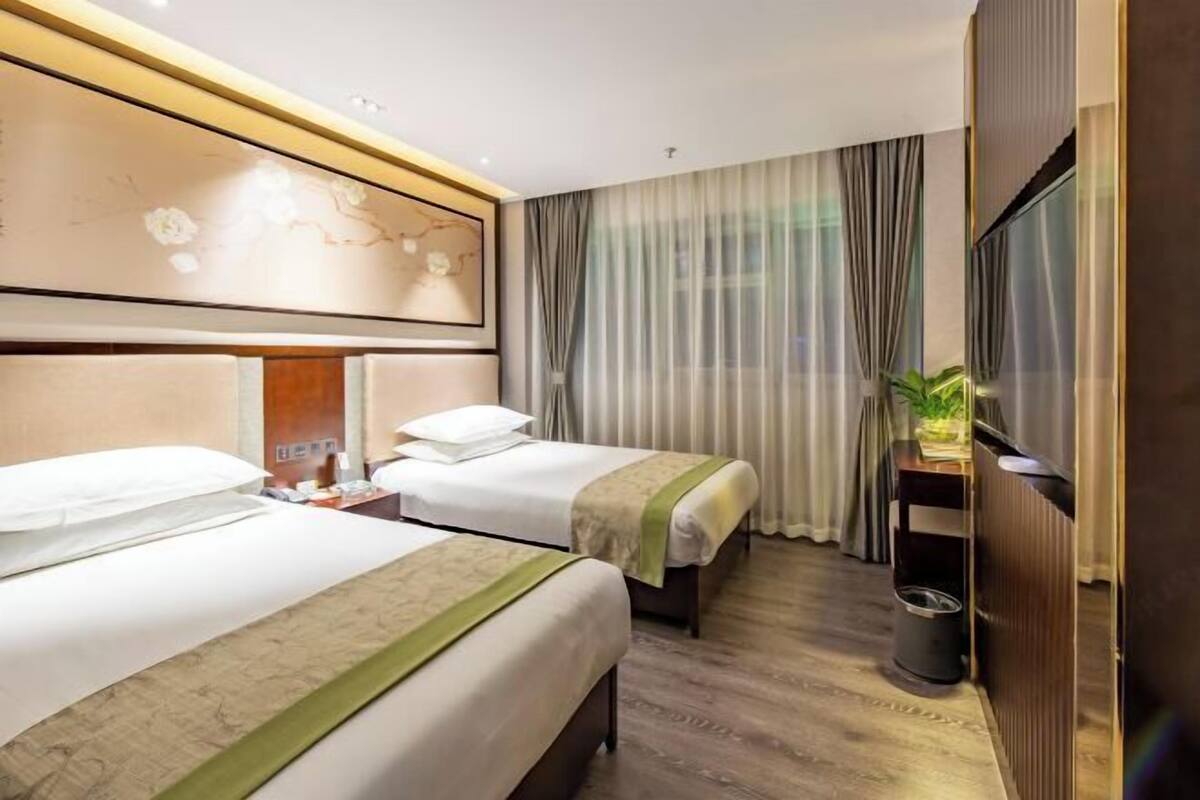 Ein Hotelzimmer mit zwei Betten, einem Fernseher, einer Pflanze und einem großen Fenster mit Vorhängen.