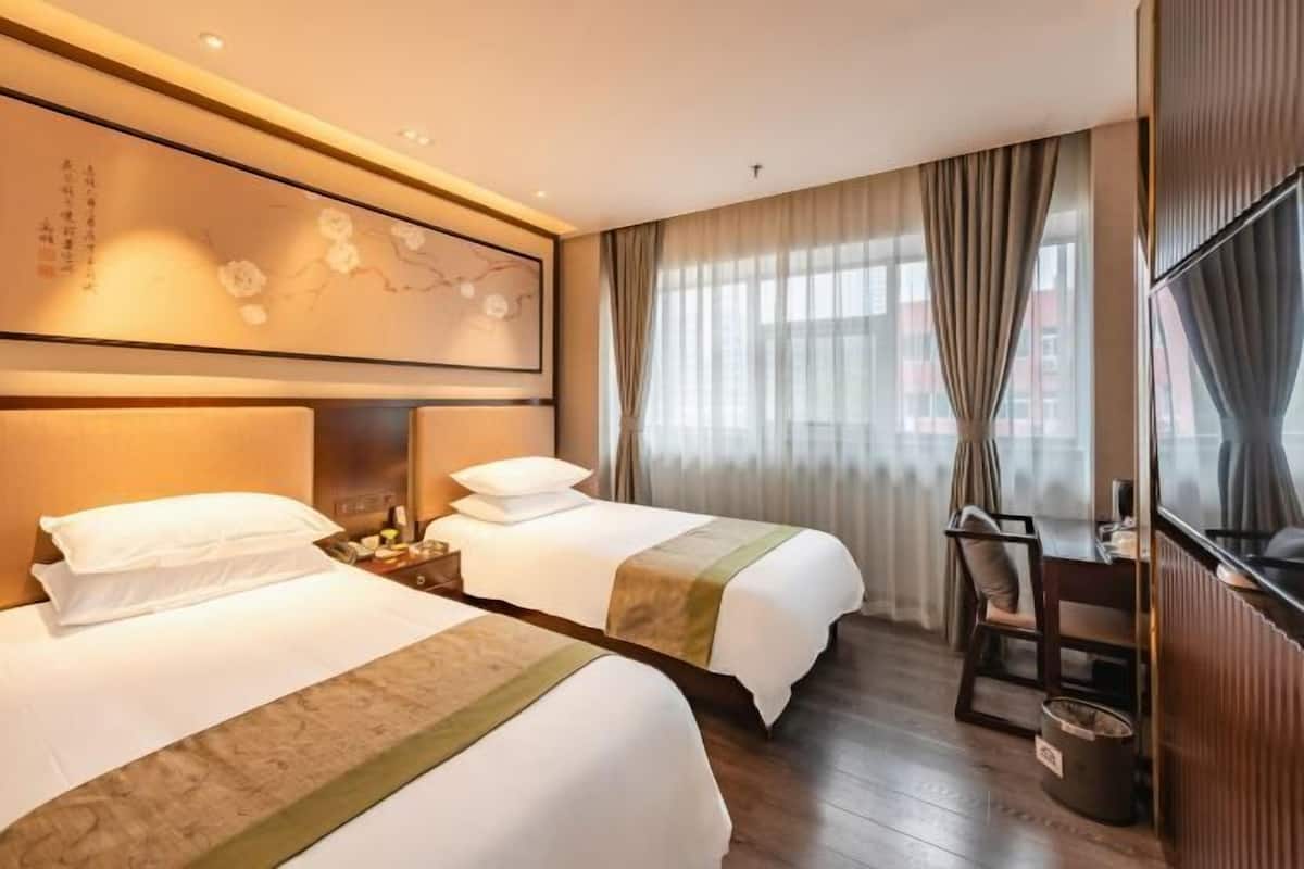 Ein Hotelzimmer mit zwei Betten, einem Schreibtisch, einem Stuhl, einem Fernseher und einem großen Fenster mit Vorhängen.