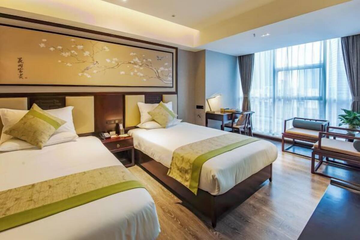 Ein Hotelzimmer mit zwei Betten, einem Schreibtisch, einem Sessel und einem großen Fenster mit Vorhängen.