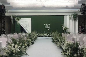 Un espace événementiel élégant, avec un décor verdoyant, des compositions florales blanches et un tapis blanc menant à une scène.