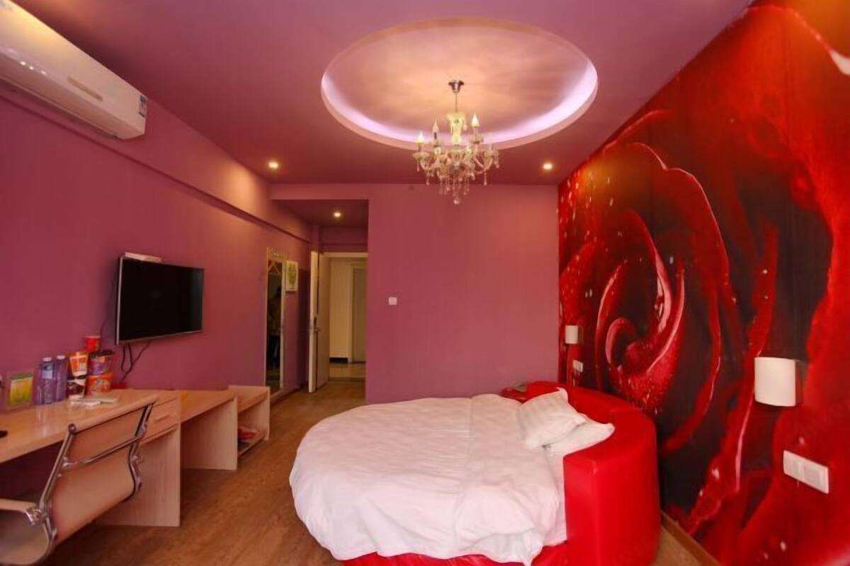 A room with a bed, a desk, a TV, and a wall with a red abstract design.
