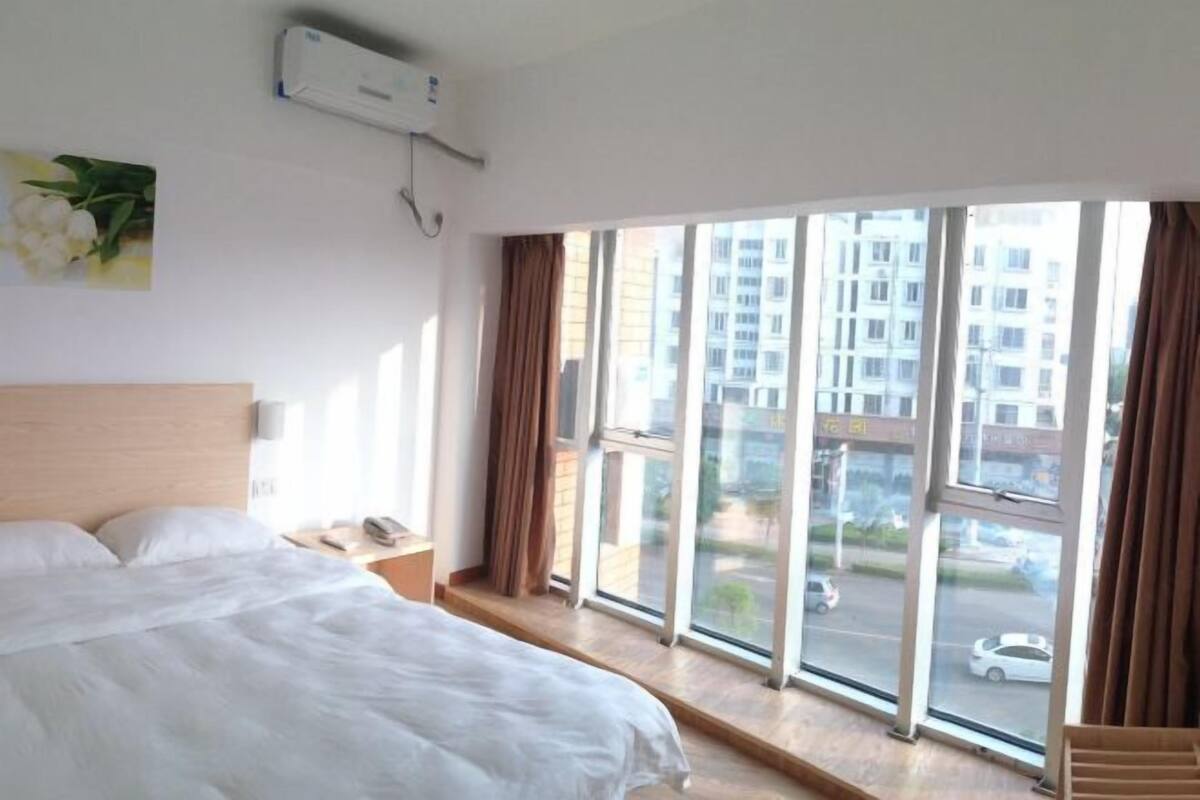 A hotel room with a large bed, a window with curtains, a wall-mounted air conditioner, and a view of the city.