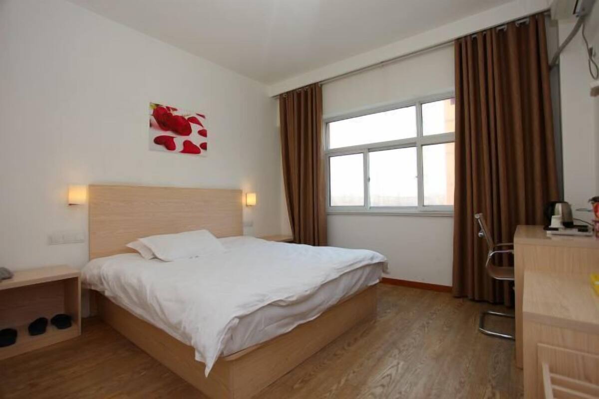 A bedroom with a wooden bed, a bedside table with a lamp, a window with curtains, and a wall-mounted artwork.
