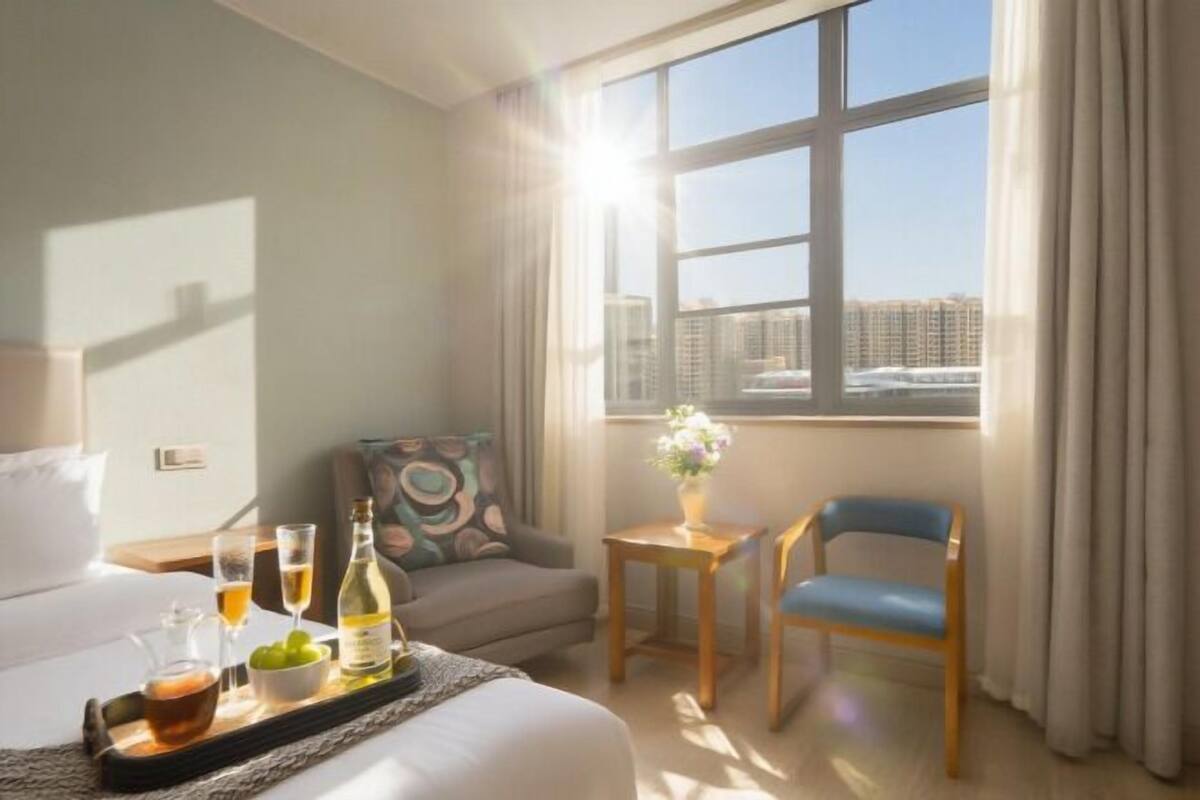Une chambre d’hôtel avec un lit, un canapé, une chaise, une petite table avec un vase et une fenêtre avec des rideaux.
