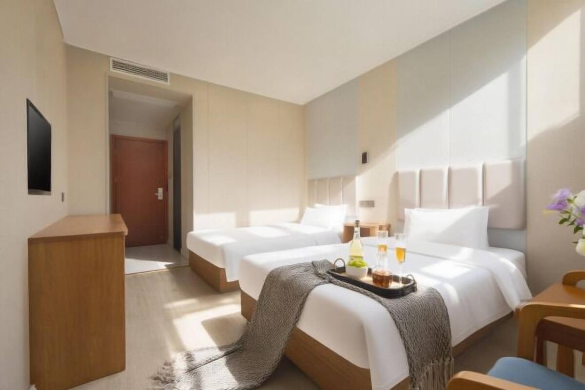 Une chambre d’hôtel avec deux lits, un bureau en bois, une chaise, une petite table sur laquelle se trouve un plateau avec divers objets, et un téléviseur fixé au mur.