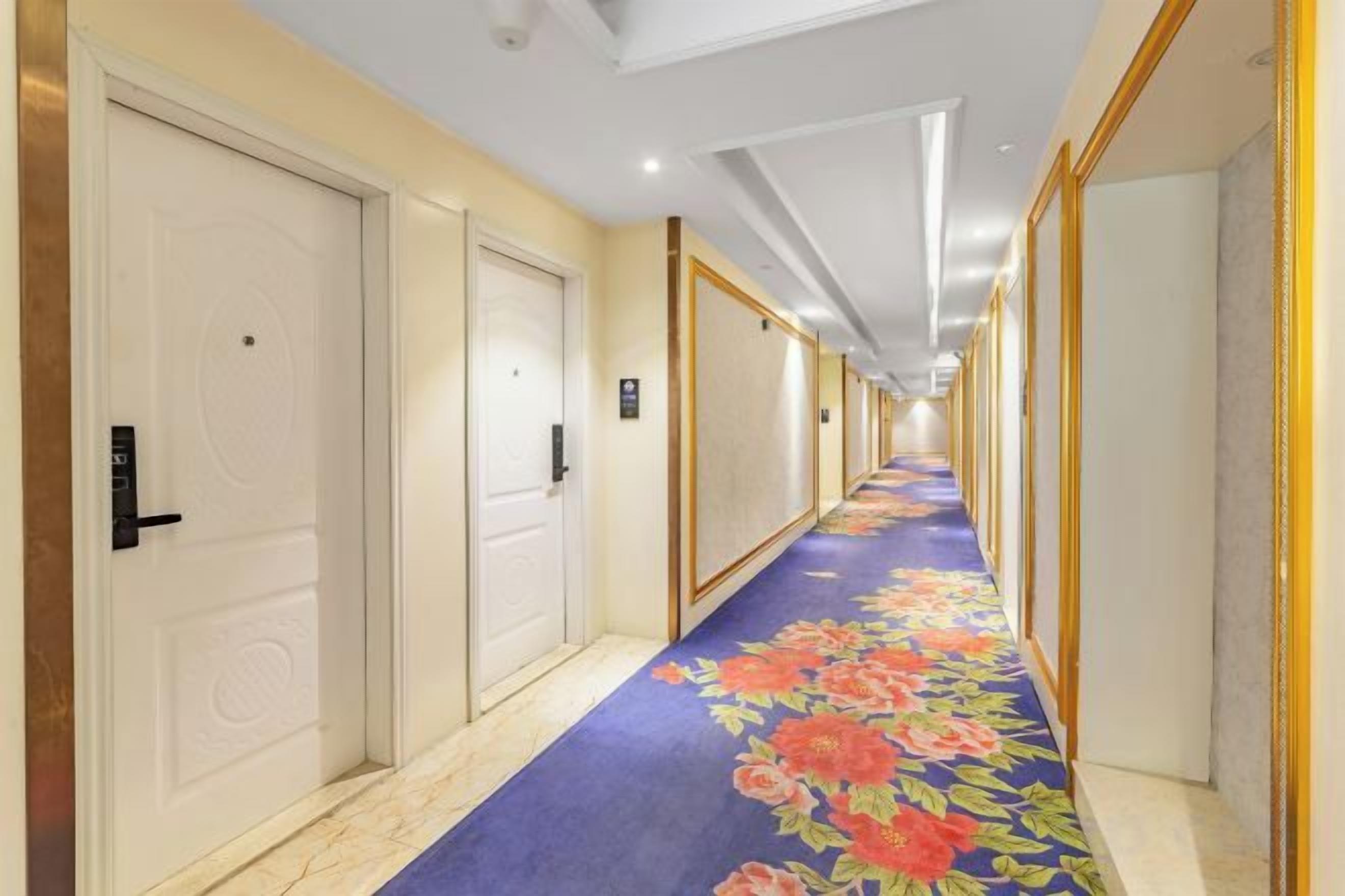 Un couloir d’hôtel avec des portes des deux côtés, une moquette à motifs floraux et un plafond doté d’un éclairage encastré.