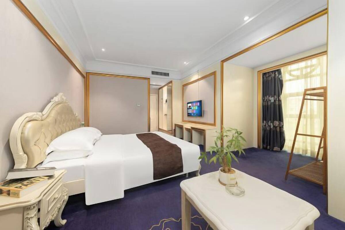 Ein Hotelzimmer mit einem großen Bett, einem Nachttisch, einem Fernseher, einem kleinen Tisch mit Pflanze und einem Fenster mit Vorhängen.