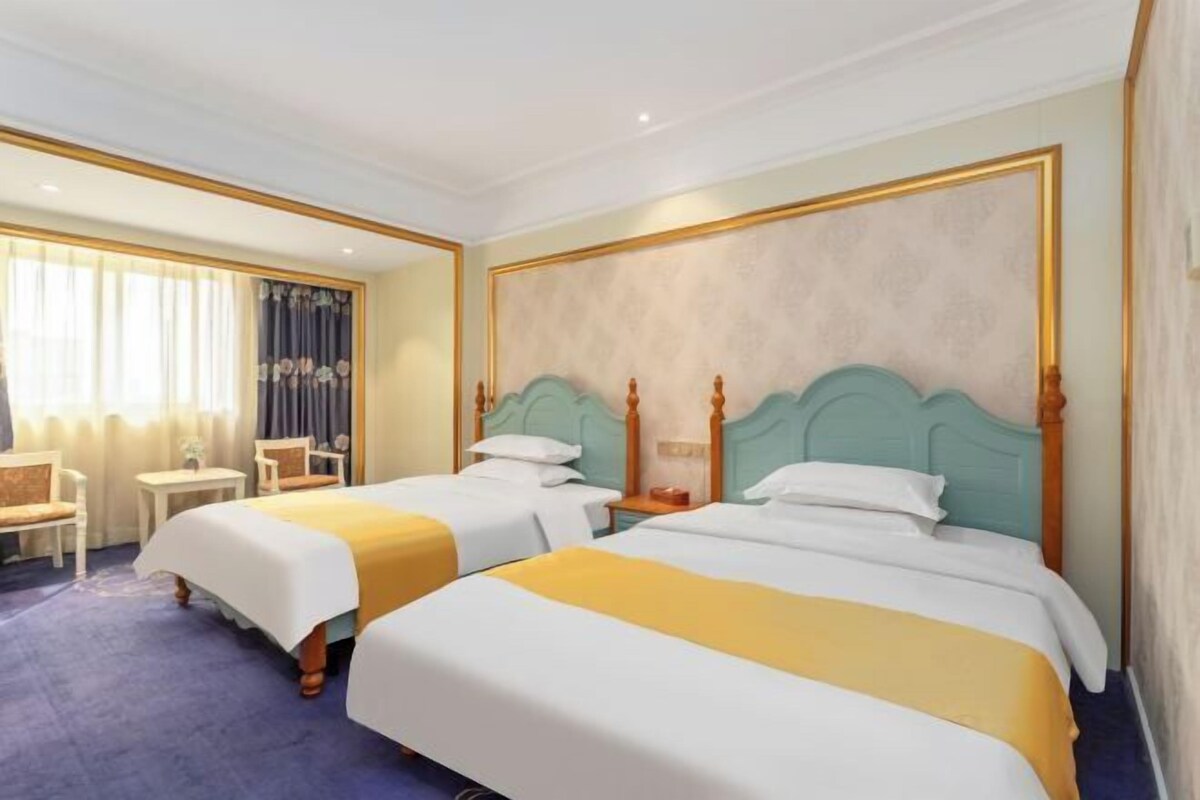 Habitación de hotel con dos camas, una mesita, una silla y una ventana con cortinas.