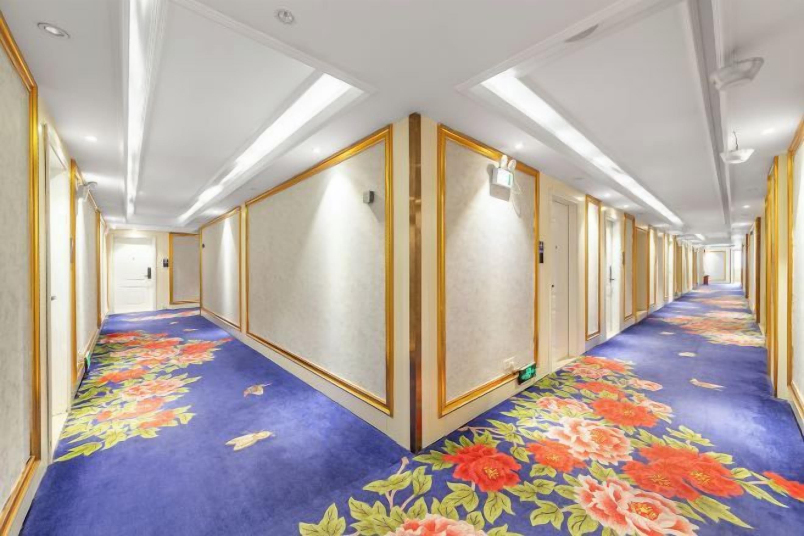 A hotel corridor with a floral carpet, doors on both sides, and overhead lighting.