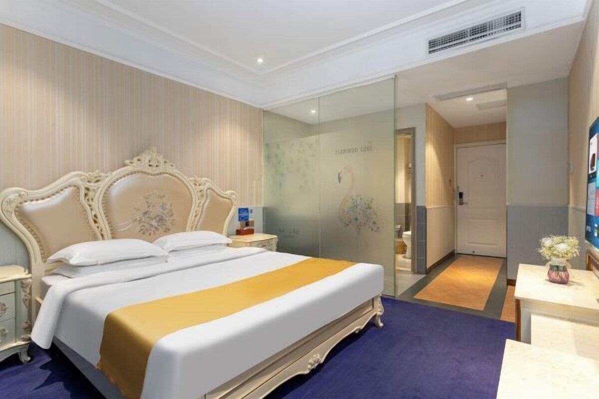 Una habitación de hotel con una cama grande, una mesita de noche y un espejo, y una puerta que da a otra habitación.