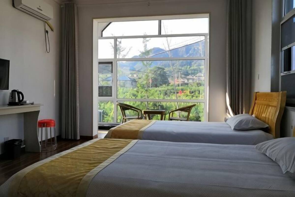 A hotel room with two beds, a desk with a kettle, a chair, and a large window with a view of greenery.