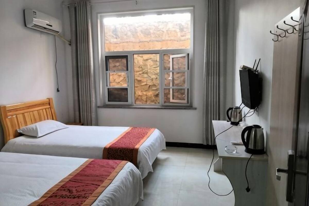 A hotel room with two beds, a window with curtains, a desk with a kettle, and a wall-mounted TV.