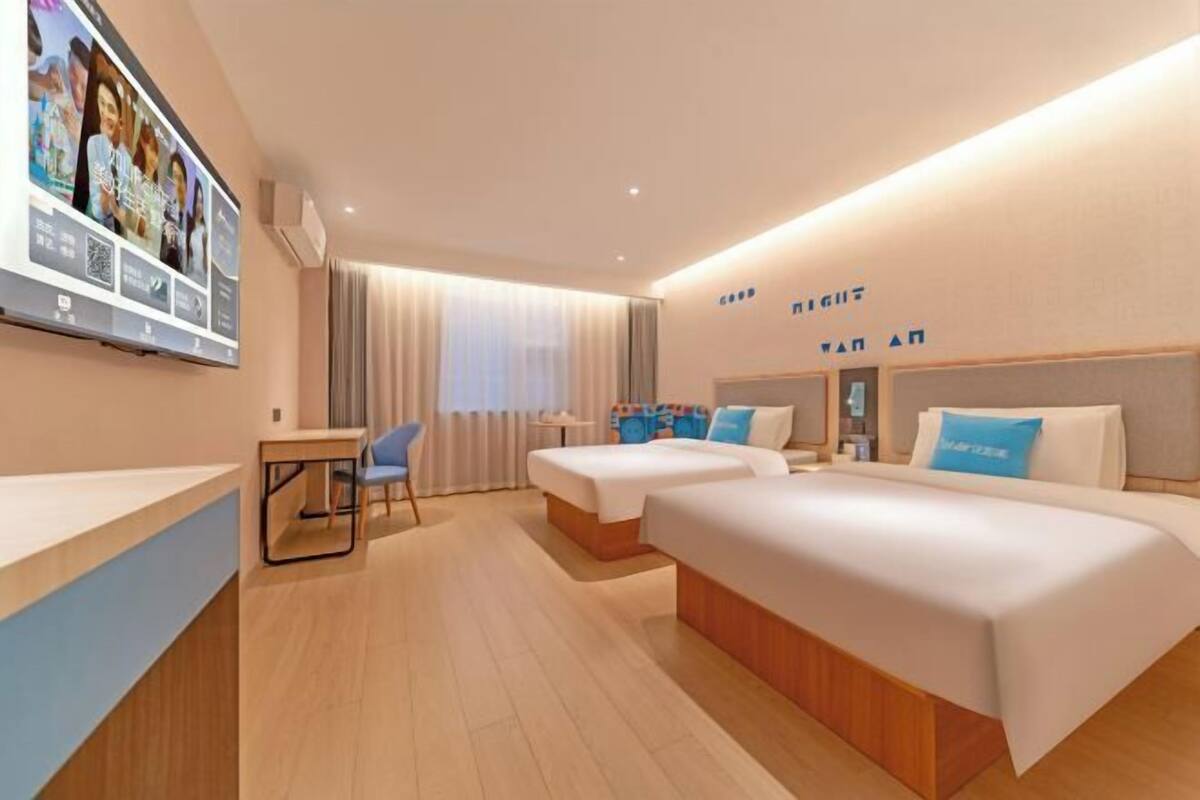 Quarto de hotel com uma televisão de tela grande, duas camas, uma escrivaninha e uma cadeira.