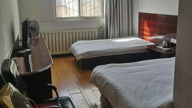 A hotel room with two beds, a desk with a chair, a radiator, and a window with curtains.