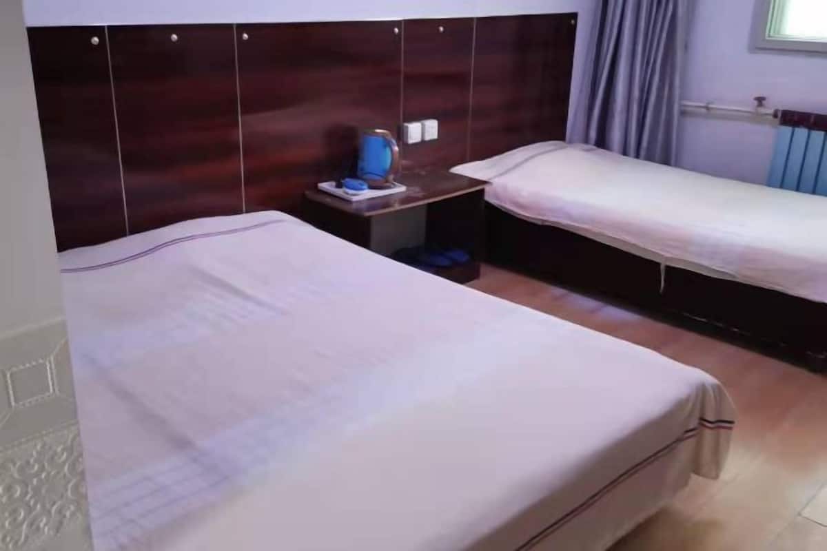Habitación de hotel con dos camas, cabecera de madera, mesita de noche con lámpara y ventana con cortinas.