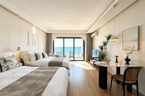 Habitación de hotel con dos camas, un escritorio, una silla, televisor y balcón con vista al mar.