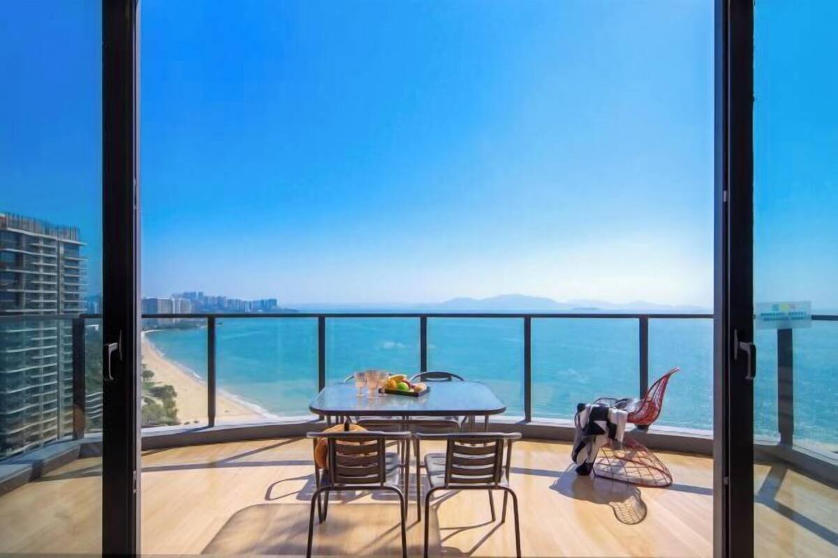 Un balcón con mesa y sillas, una silla roja, y vista al mar y edificios a lo lejos.