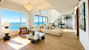 Een ruime woonkamer met een groot raam met uitzicht op zee, een moderne bank, een salontafel en een trap naar een hoger gelegen verdieping.