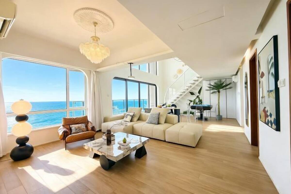 Un espacio habitable amplio con un gran ventanal con vistas al mar, un sofá moderno, una mesa de centro y una escalera que conduce a un nivel superior.