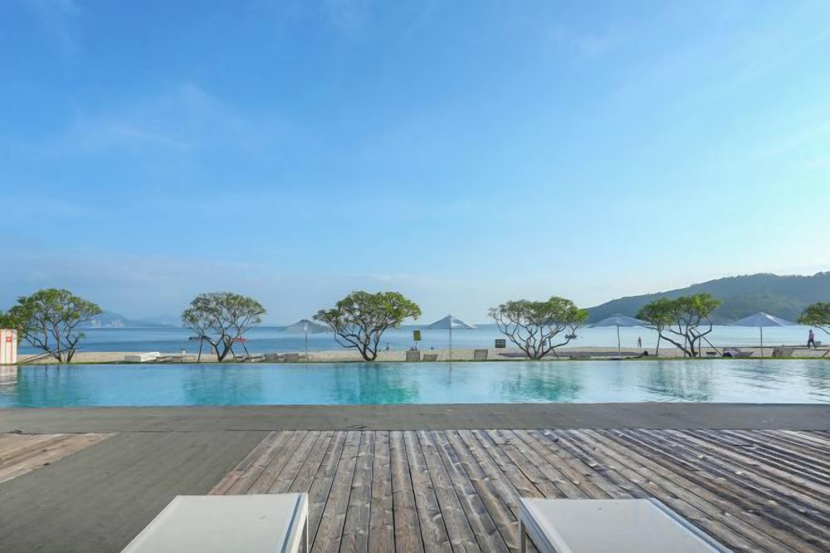 Ein Poolbereich mit Holzterrasse, Liegestühlen und Blick auf das Meer und die entfernten Hügel.
