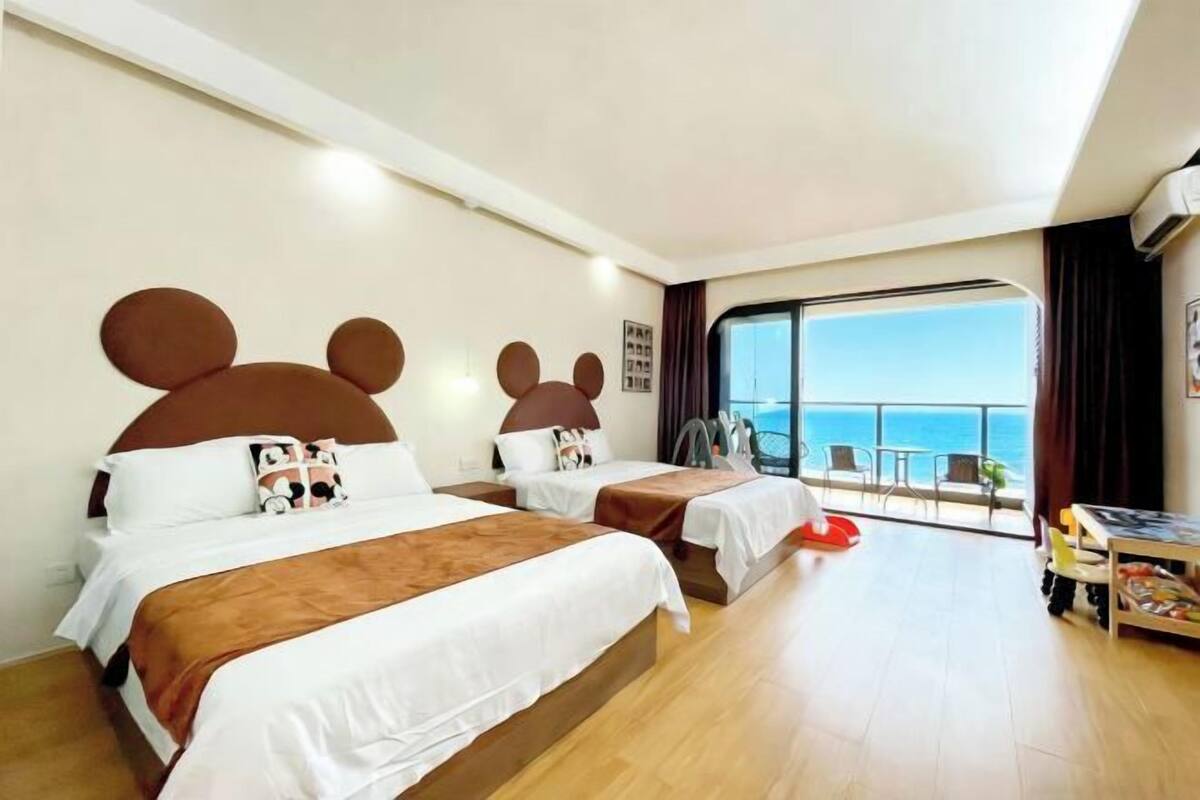 Habitación de hotel con dos camas, cada una con cabeceros de Mickey Mouse, un balcón con vistas al mar y una pequeña mesa con una silla.
