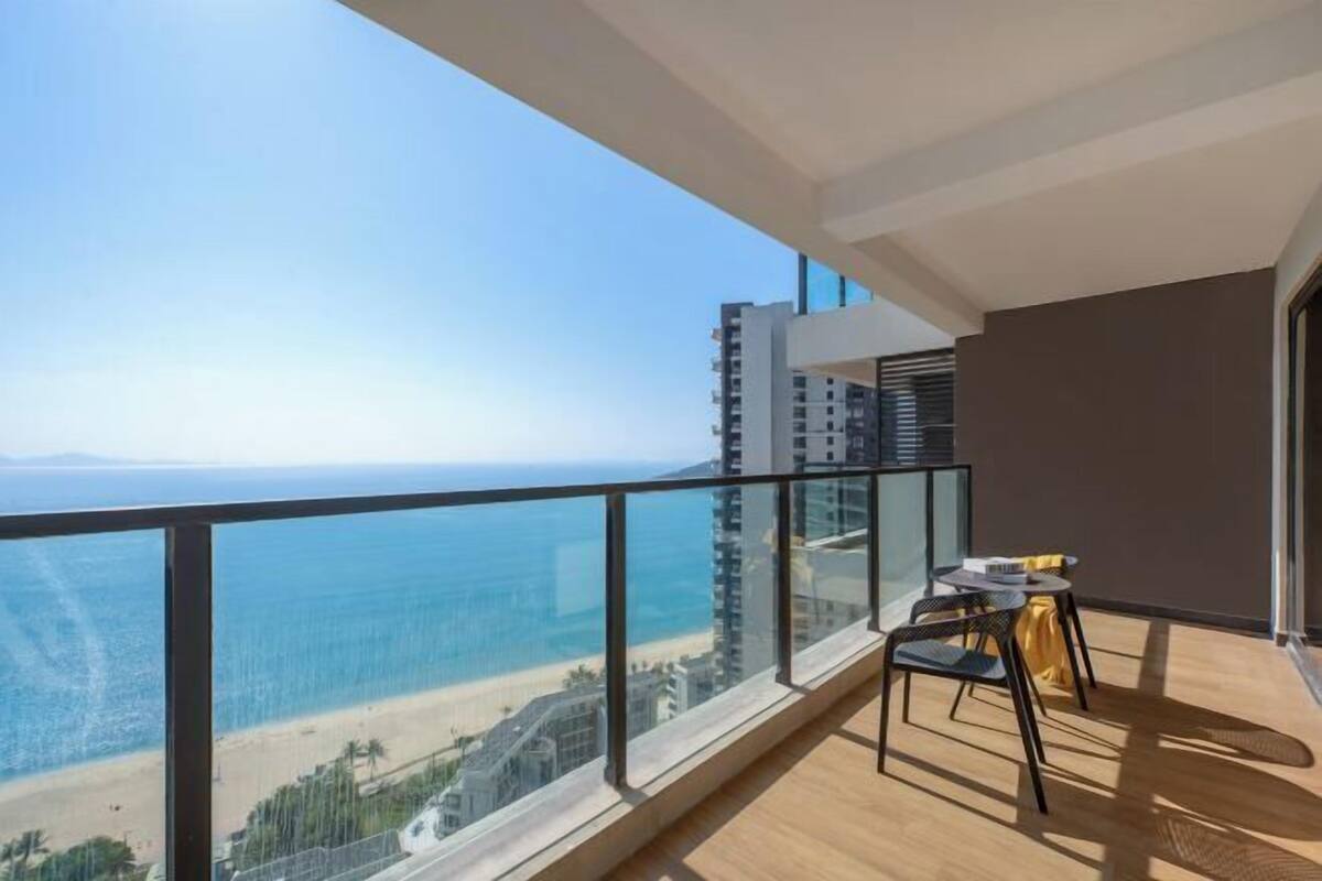 Un balcón con vista a la playa y al océano, una pequeña mesa con sillas y una barandilla de vidrio.