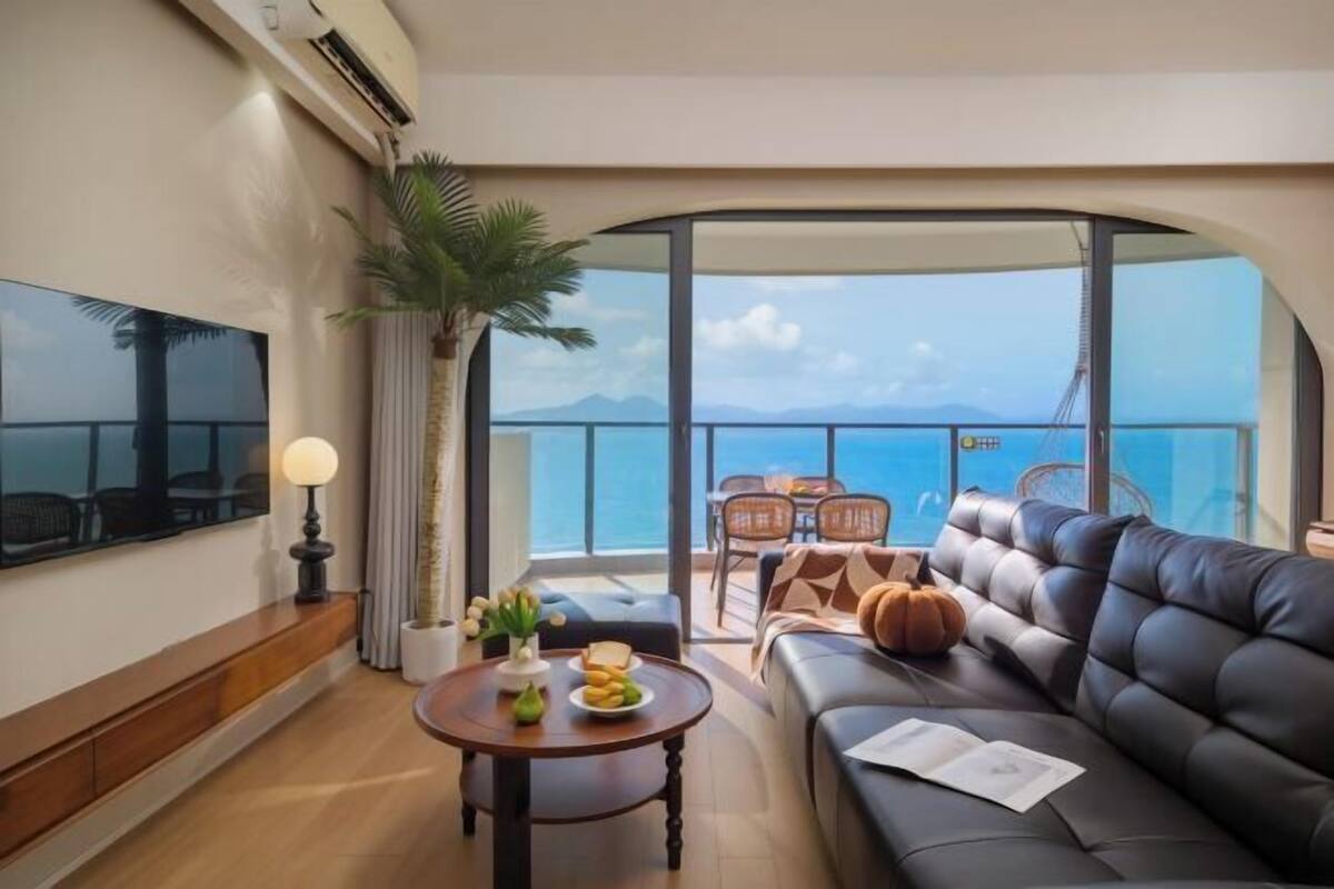 Una sala moderna con sofá, una mesa de centro y televisor. Cuenta con un balcón con vistas al mar y a las montañas.