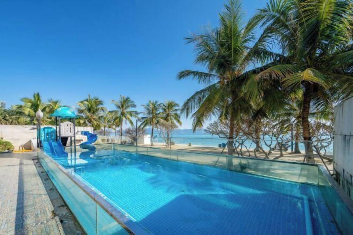 Una piscina azul cristalina con tobogán, rodeada de palmeras y vista al mar.