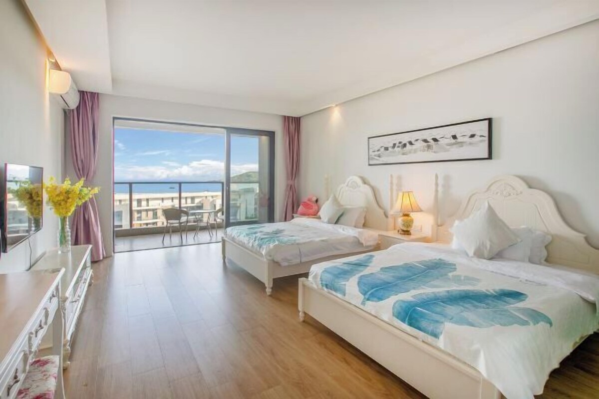 Habitació d'hotel amb dos llits, un balcó que dona a una vista panoràmica i una taula de treball amb un ram de flors.