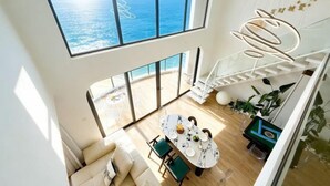 Een ruime kamer met een groot raam met uitzicht op de oceaan, een eethoek met een ronde tafel, en een trap die naar een hoger gelegen niveau leidt.