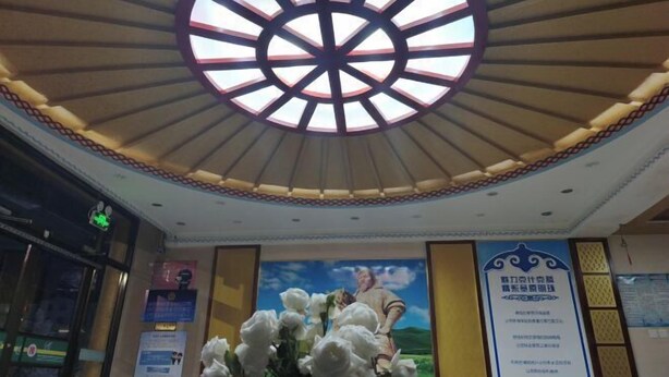 A lobby with a large circular skylight, a poster of a man and woman, and a vertical banner with text. - Prairie Pearl Express Hotel (Hexigten Banner)