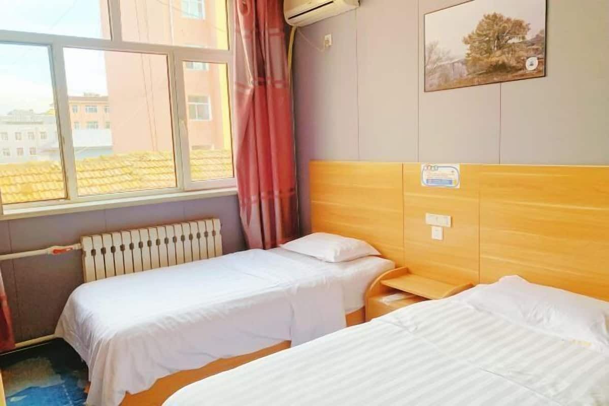 A hotel room with two single beds, a window with a view of buildings, an air conditioning unit, and a painting on the wall.