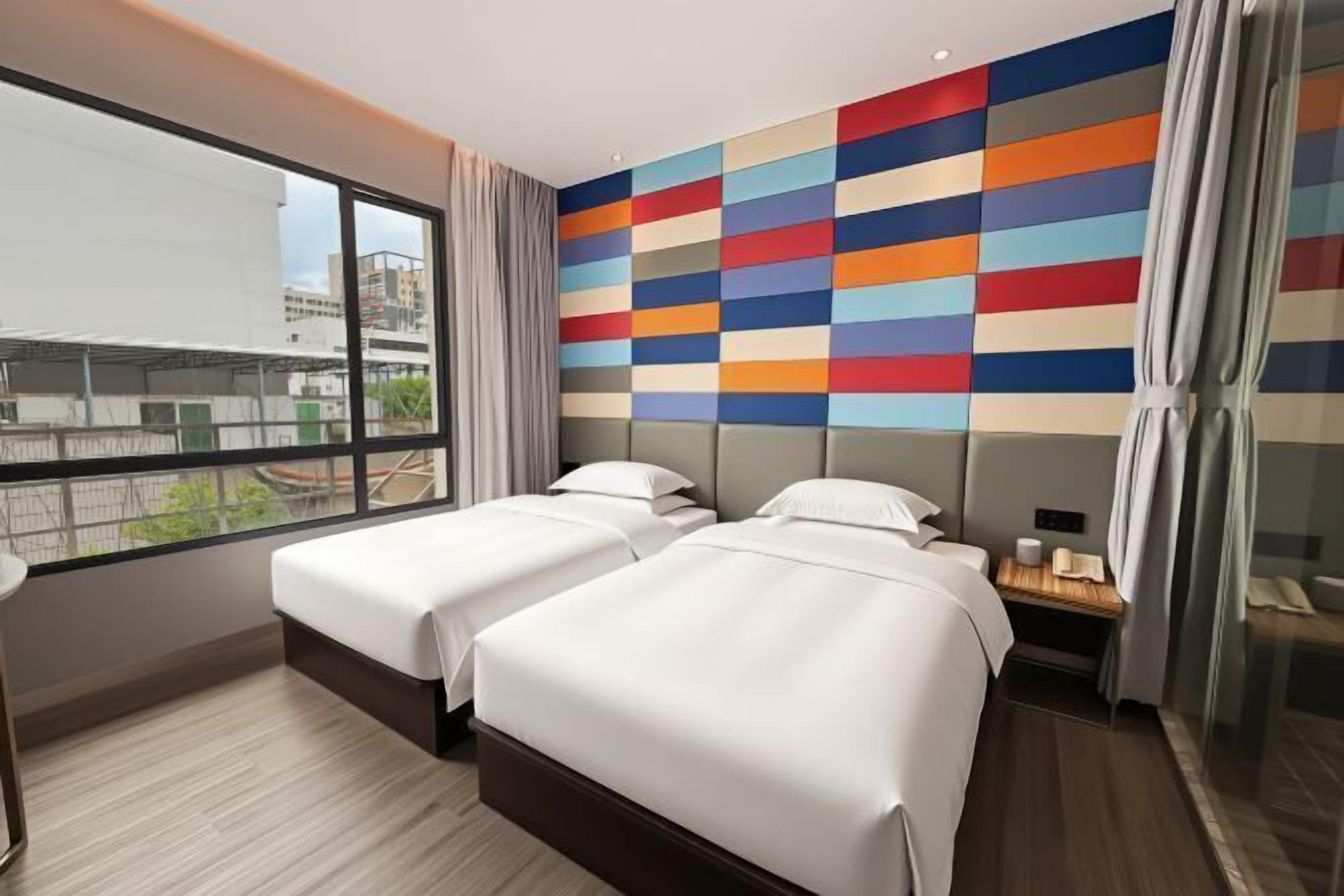 A hotel room with two beds, a large window, and a colorful wall design.