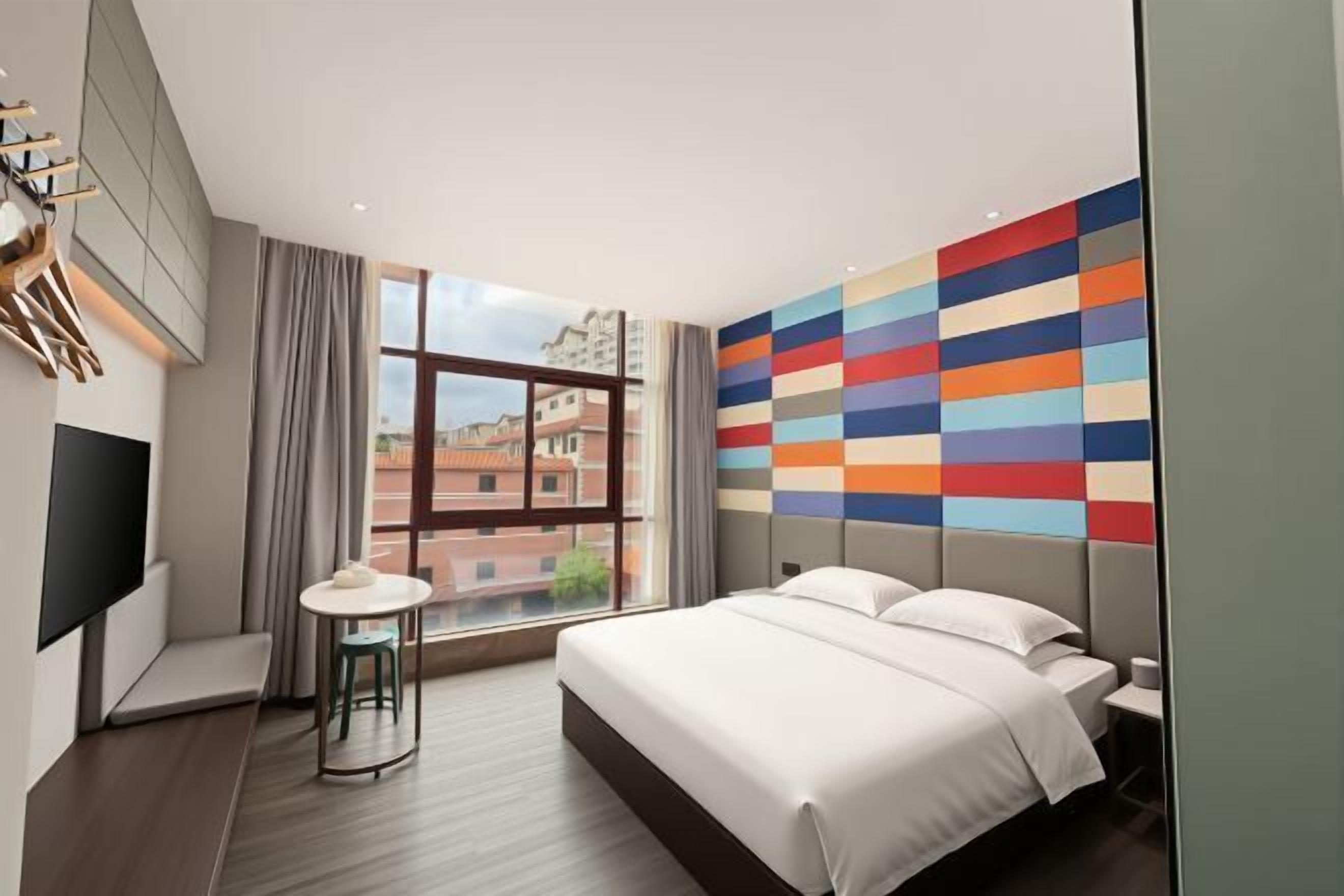 A modern hotel room with a large bed, a small round table, a TV, and a large window with a city view.
