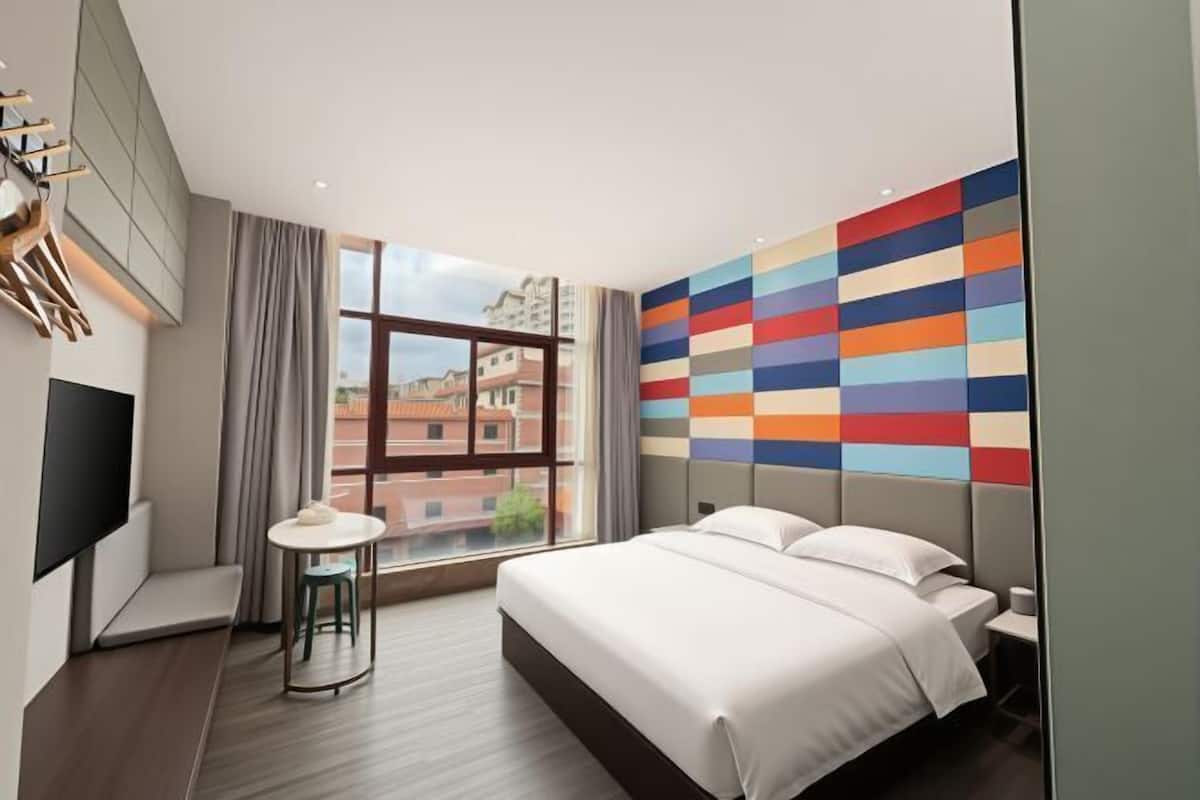 Una habitación de hotel moderna con una cama grande, una pequeña mesa redonda, un televisor y un ventanal con vista a la ciudad.