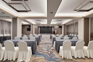 Un salón de conferencias con filas de mesas y sillas, un escenario y suelo alfombrado.