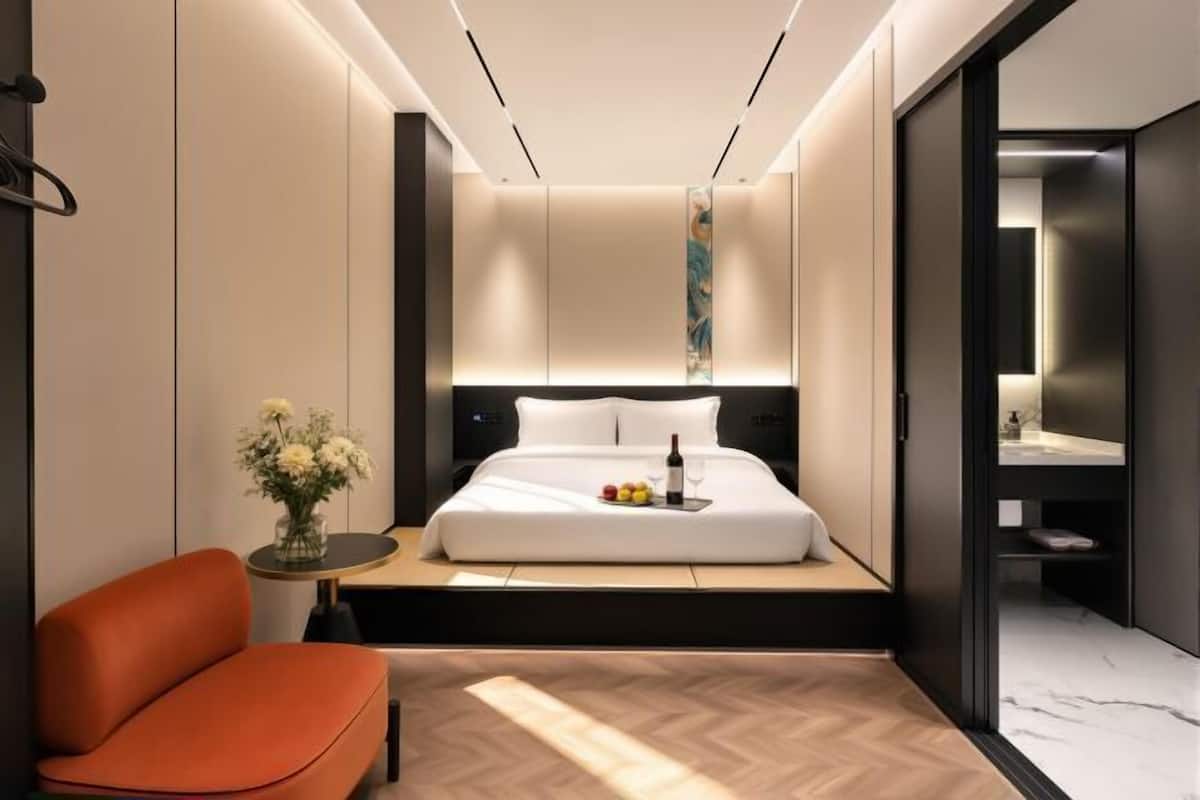 Una habitación de hotel moderna con una cama, una mesita con flores, una silla y un baño visible a través de una puerta abierta.